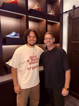 Two men posing together in what appears to be a sneaker shop; the man on the left wears a shirt reading 'i give a blank about crypto' and the man on the right wears a black t-shirt and glasses.