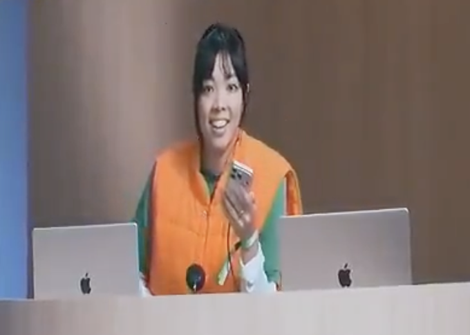 A woman in an orange vest stands behind a desk with two Apple laptops, holding a smartphone and appearing to speak.