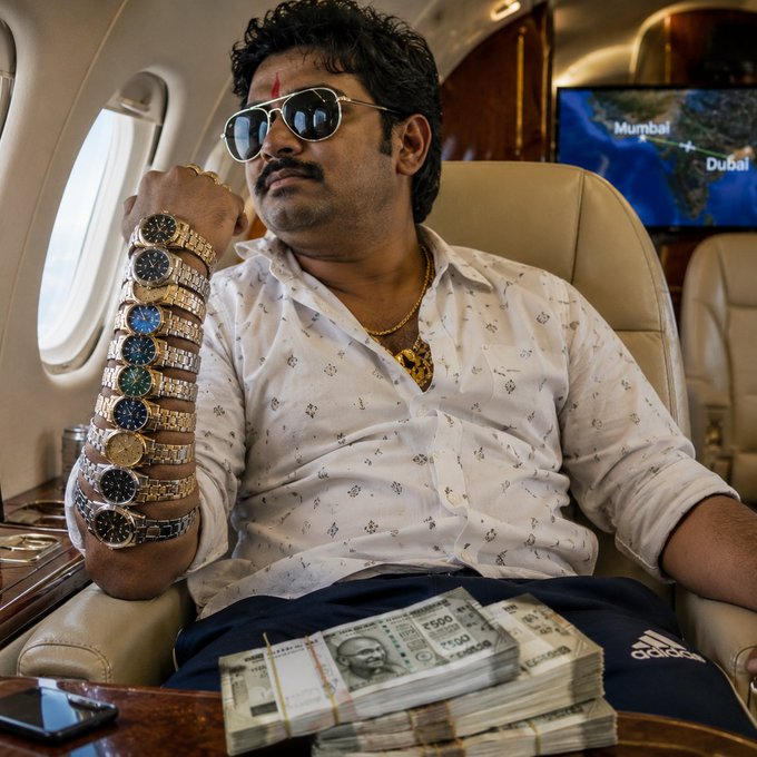 A man wearing sunglasses and an elaborate stack of luxury watches sits inside a private jet with a flight path displayed from Mumbai to Dubai, surrounded by stacks of Indian rupee notes. The image satirically visualizes the "pump gains" brag of the post, using exaggerated wealth symbols—a private jet, designer timepieces, and cash—to mock crypto pump-and-dump culture and the "stay long" rallying cry.
