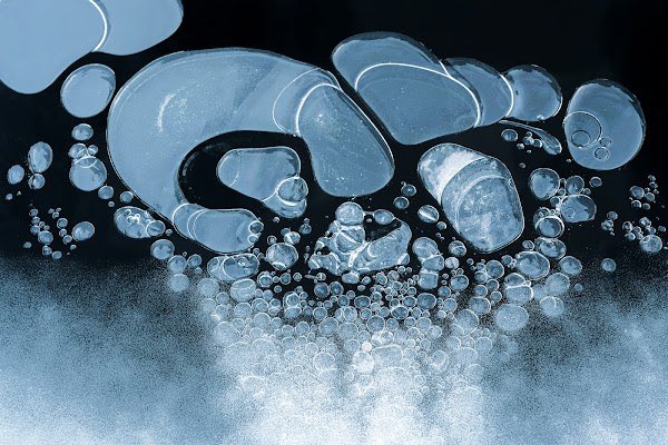 An abstract, high-contrast macro photograph showing translucent blue bubbles and fluid shapes against a black background.