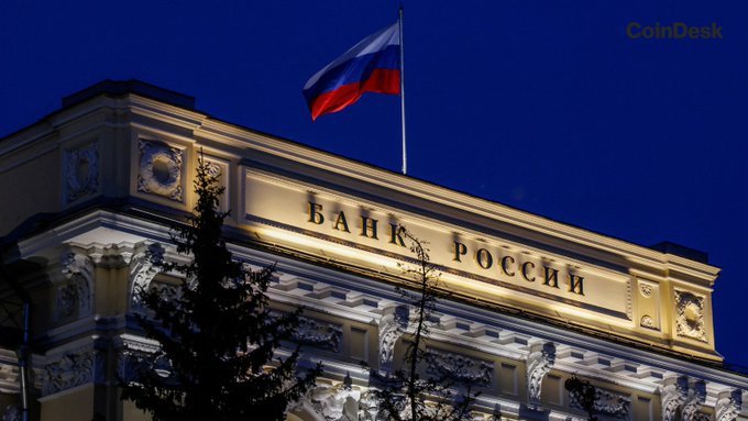 The Bank of Russia building is illuminated at night with the Russian flag flying above, displaying the Cyrillic inscription "Банк России" (Bank of Russia). The image visually anchors the news about Russia's central bank restricting retail crypto trading to only $BTC, $ETH, and $USDT on regulated exchanges.