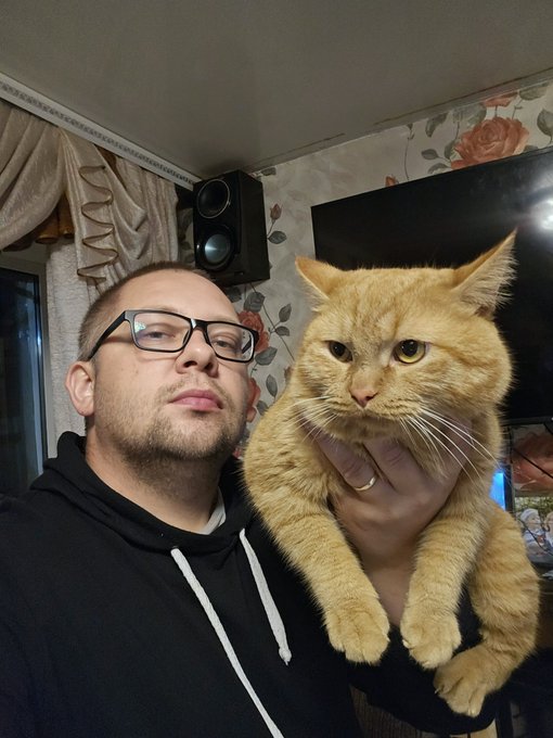 A man wearing glasses and a black hoodie takes a selfie while holding an orange tabby cat with a serious expression.