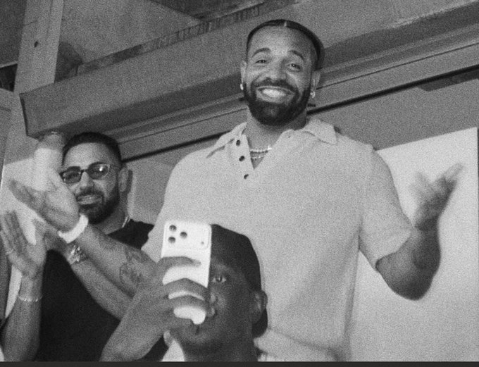 A black-and-white photo of a smiling bearded man in a white polo shirt standing with arms outstretched, surrounded by people taking photos with smartphones at what appears to be a club or event venue.