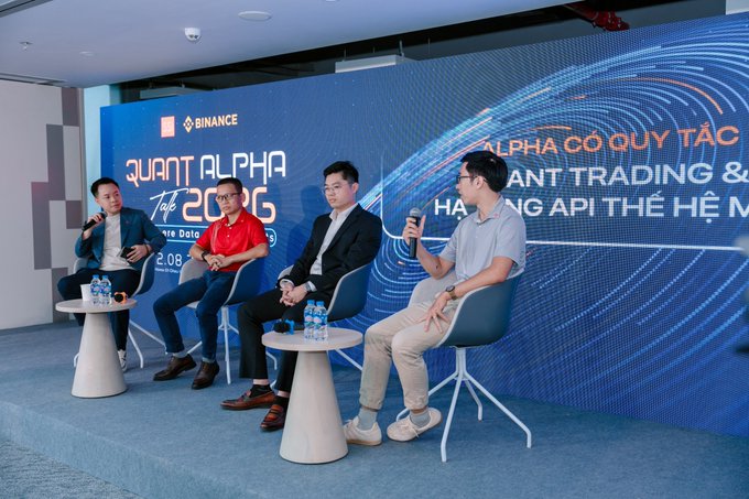 Four men sit on stage at a Binance 'Quant Alpha' event, with the backdrop displaying text about 'QUANT TRADING & HÀNG API THẾ HỆ MỚI' in Vietnamese.