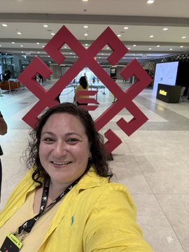 A smiling woman in a yellow shirt takes a selfie in front of a large red decorative structure in what appears to be a shopping mall or public venue.
