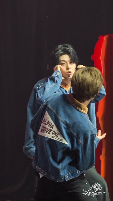 Two performers in matching blue denim jackets pose together on a dimly lit stage, with the jacket reading 'ALPHA DRIVE ONE'; watermark 'Lanjun' appears in the corner.