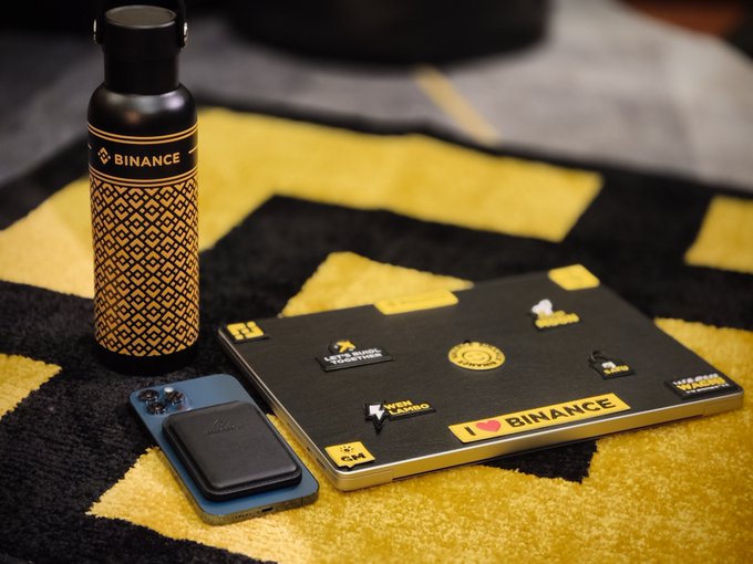 A black and yellow patterned 'BINANCE' water bottle sits on a rug next to a laptop covered in Binance-themed stickers including 'I ❤ BINANCE' and a phone with a MagSafe charger.