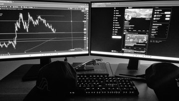 A black and white image showing a person's hands on a keyboard with two computer monitors displaying trading charts and YouTube videos in the background.