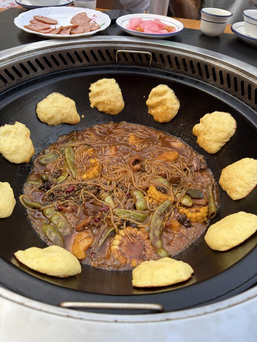 A large round grill pan contains a stir-fried noodle dish with vegetables, meat, and corn in the center, surrounded by golden fried dough or dumplings arranged around the edge, with plates of sliced sausage and pickles visible in the background.