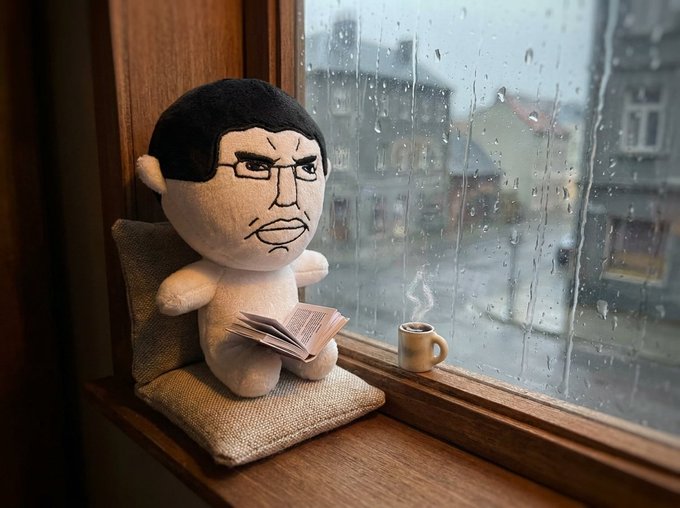 A plush toy figure with drawn-on black hair and glasses sits on a windowsill reading a book next to a steaming coffee cup, with rain streaking the window overlooking a city street.