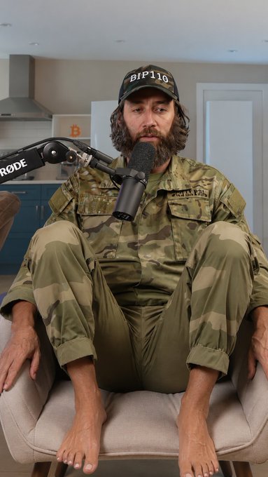 A bearded man wearing a "BIP110" cap and camouflage clothing sits barefoot in a chair with his legs pulled up, speaking into a RØDE microphone in a kitchen setting—his awkward seated pose humorously reflecting the post's joke about "increasingly creative positions" used to promote BIP110.