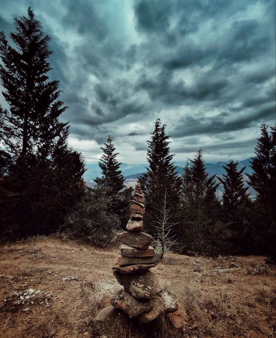 A stone cairn stands atop a dirt mound surrounded by evergreen trees under a dramatic, cloudy gray sky with mountains visible in the distance.