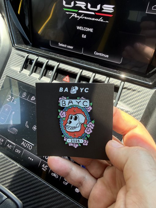 A hand holds a black 'BAYC 2026' sticker with a skull and floral design in front of a Lamborghini Urus Performante dashboard displaying 'WELCOME Ed'.