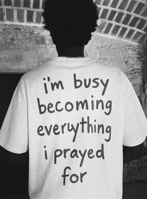 A black-and-white photo of a person viewed from behind, wearing a t-shirt with the motivational text 'i'm busy becoming everything i prayed for'.