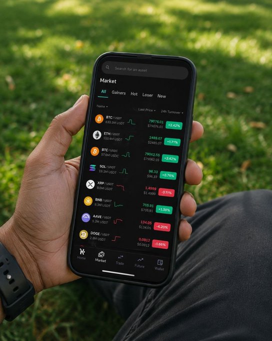 A hand holds a smartphone displaying a cryptocurrency market app against a grassy background, showing various tokens including BTC at $79016.01 (+2.42%), ETH, SOL, XRP, BNB, AAVE, and DOGE with their respective prices and 24-hour gains or losses in green and red indicators. The market overview ties directly to the post by inviting viewers to scan the listed tokens and pick one that stands out this August.
