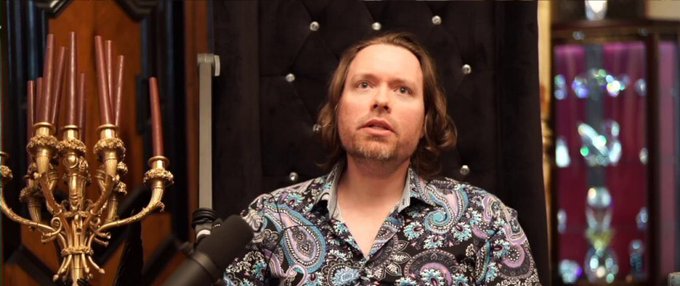 A man with shoulder-length brown hair wearing a paisley patterned shirt, sitting in front of a black tufted backdrop with a gold candelabra visible.