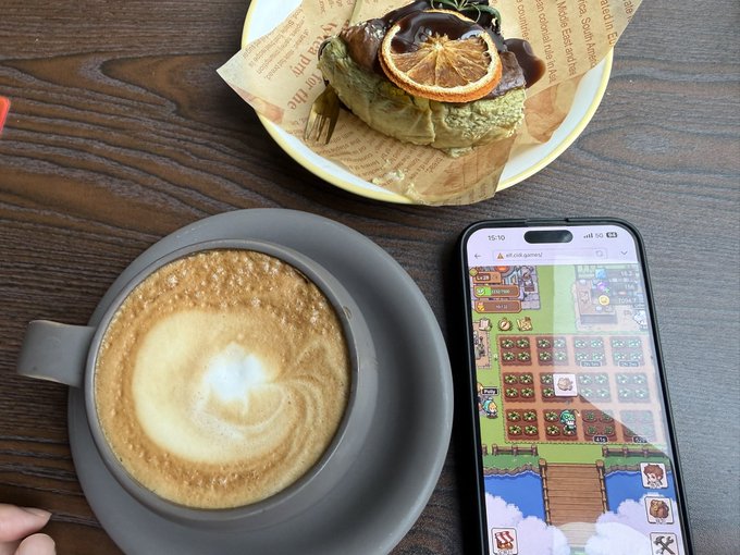 A latte with foam art, a citrus-topped pastry, and a smartphone playing "Growscape" sit on a wooden table during a relaxing café break, offering a well-deserved pause from farming water radish in-game.