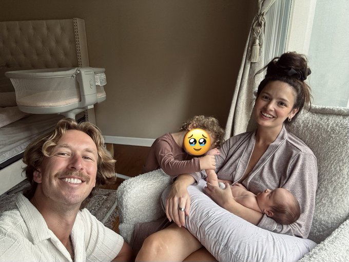 A family selfie showing a smiling man with a mustache, a woman with dark hair in a bun holding a swaddled newborn, and a young child with curly hair (whose face is covered by a smiley emoji sticker) sitting on a fluffy chair.