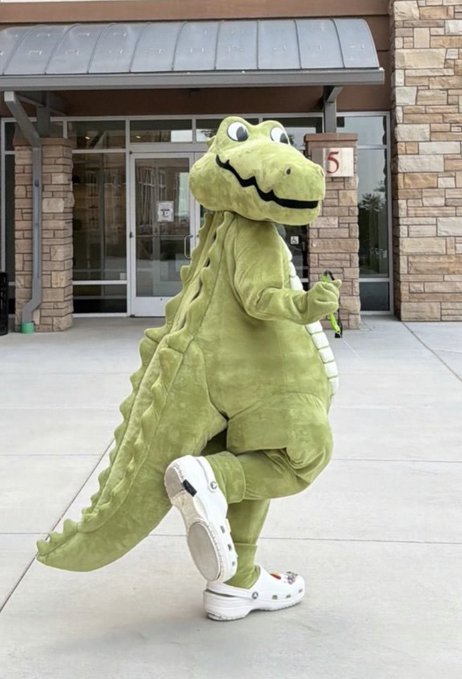A person wearing a green dinosaur or crocodile costume and white Crocs walks outside a store.