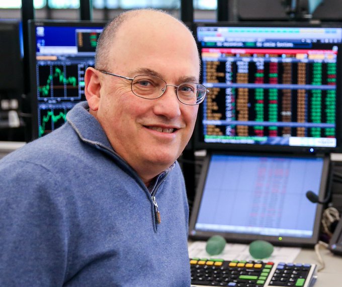 A smiling, bald man wearing glasses and a blue quarter-zip sweater poses in front of multiple computer monitors displaying financial trading charts, graphs, and data screens.