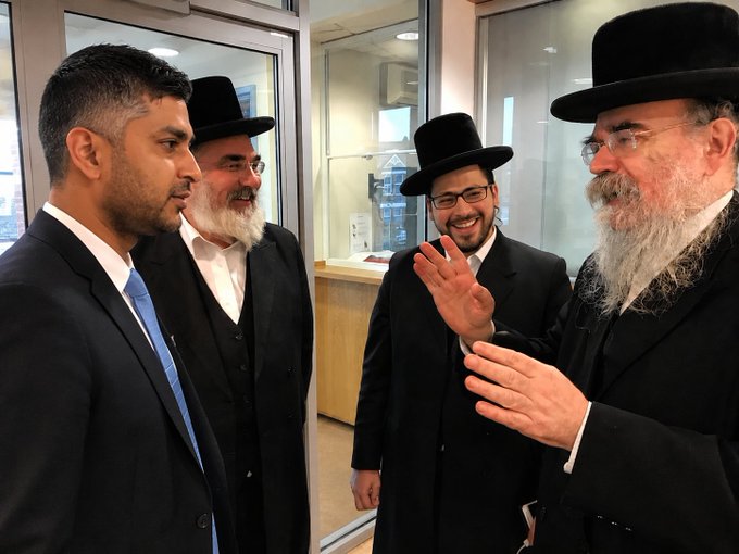Four men in dark suits and black hats, including one with a long white beard and another wearing glasses, converse and gesture indoors.