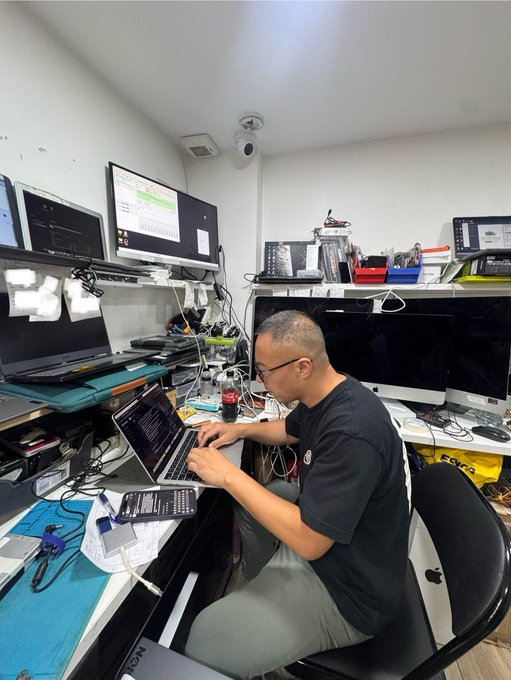 A candid photo of a man in a black t-shirt and glasses working on a laptop at a cluttered multi-monitor workstation in a small room. The desk is filled with computer monitors, cables, a keyboard, and electronic equipment.