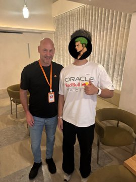 Two men pose together at what appears to be an indoor event; one wears an orange lanyard while the other sports a green visor and an "Oracle Red Bull Racing" t-shirt. The casual, friendly setting aligns with the MetaMask dinner in Singapore mentioned in the caption, capturing a moment of networking among the international attendees.