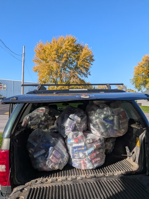 The open rear of a vehicle is packed with clear plastic bags filled with aluminum cans, parked outdoors beneath a blue sky and autumn-colored trees.