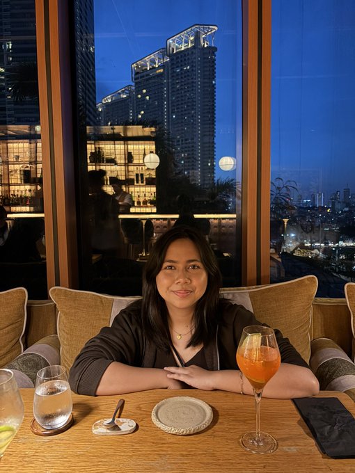 A woman smiling at a restaurant table with an orange cocktail, seated by a window overlooking illuminated skyscrapers at dusk.