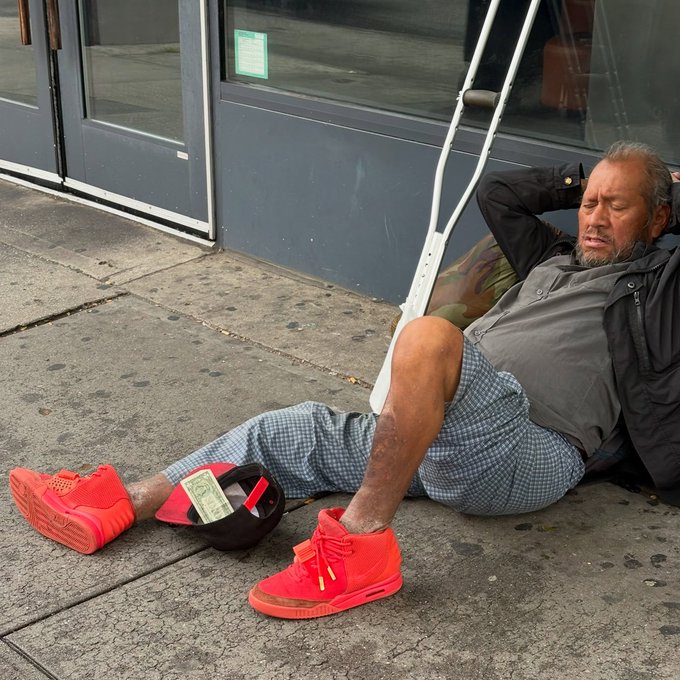 A bearded man rests on a city sidewalk with his head against crutches, wearing a dark jacket, plaid shorts, and bright red sneakers, while a baseball cap containing cash sits in front of him as an open donation cup.