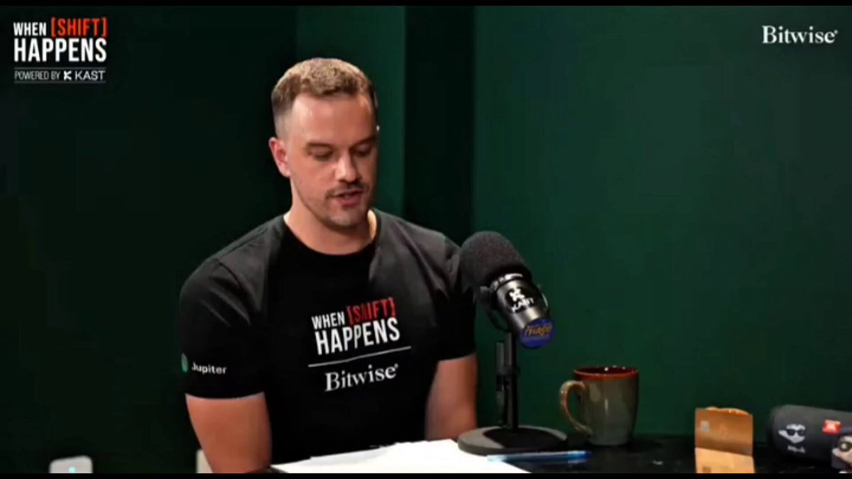 The image shows a man sitting at a table with a microphone, wearing a black t-shirt that says "When It Happens" on it. The background of the image features a green wall, and there are two cups placed on the table in front of him - one is a coffee cup, while the other appears to be a water bottle. The post discussing the launch of Kalshi's product, which was influenced by the concept of "hydration" or "hydrogen peroxide", seems to be related to this scene as it might discuss how the company's founder came up with the idea for their product and its connection to the hydration trend.