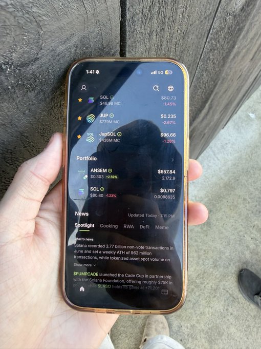 A hand holds a smartphone displaying a crypto portfolio tracker, showing holdings including ANSEM ($0.303, +2.59%, valued at $657.84) and SOL ($80.80, down 1.23%), alongside Solana-related news. The image illustrates the creator's excitement of seeing the $ANSEM token airdropped into their real-life crypto wallet.