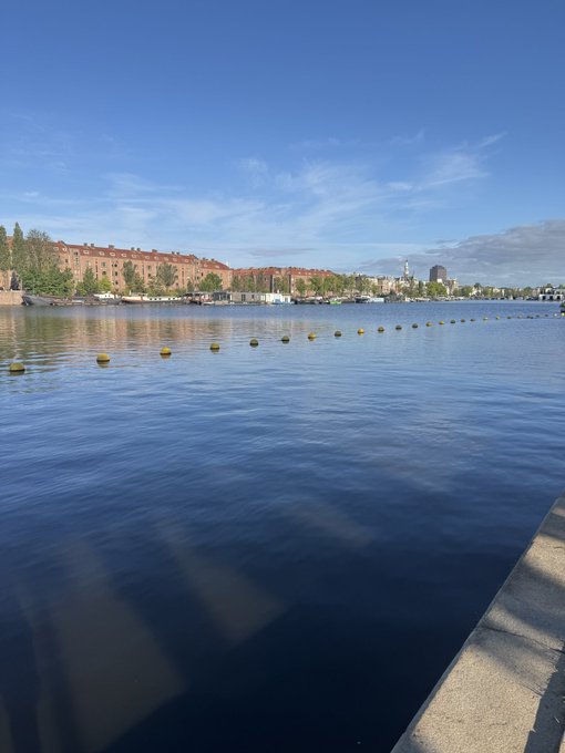 A calm waterfront scene at sunrise features a line of yellow buoys stretching across still water, with a row of red-roofed buildings and distant city skyline visible on the opposite shore under a clear blue sky—capturing the peaceful start to a "Good morning!"