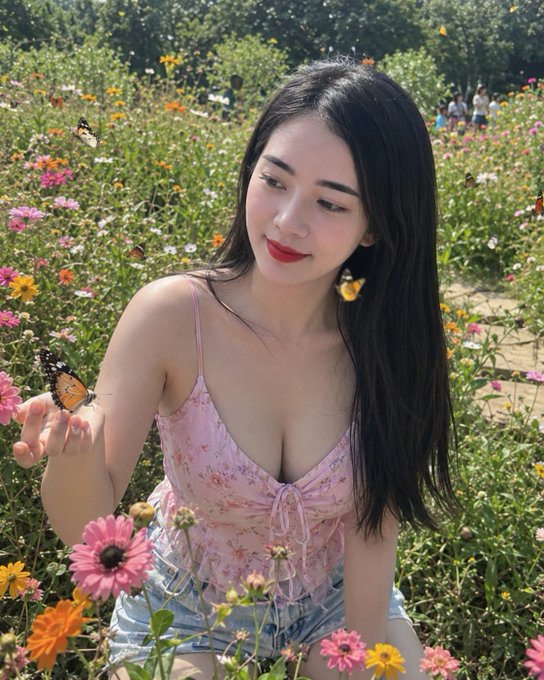 The image features a woman sitting among flowers with long hair wearing a pink top. She has her arms around herself, possibly posing for a photo or enjoying the scenery. The background includes various types of colorful flowers that create a vibrant atmosphere. There is also an element of technology in the scene, as there are multiple butterflies flying around and landing on different flowers. This suggests that the woman might be at a park or garden where people can enjoy nature while still being connected to modern technology through their smartphones.