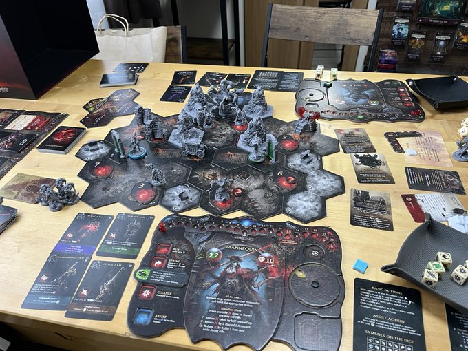 An overhead view of a board game in progress on a wooden table, featuring hex-based terrain tiles, miniature figures, cards, dice, and player boards for what appears to be a dungeon-crawler game.