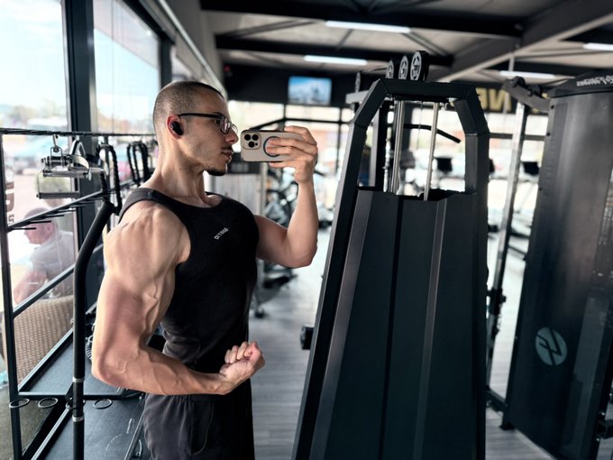 The image features a man wearing glasses, black tank top, and shorts who appears to be flexing his muscles at a gym. He is holding a phone up to take a picture of himself while working out with weights. The photo has been edited to make him look like he's in the middle of an intense workout session.