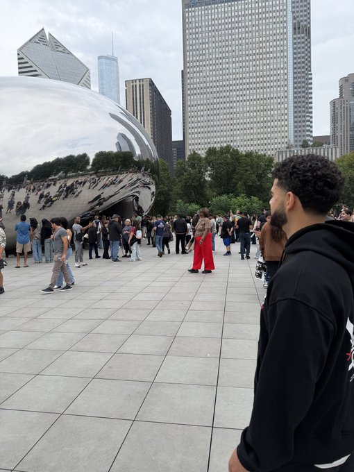 A man with curly hair wearing a black hoodie stands at Chicago's Millennium Park, looking toward the Cloud Gate (Bean) sculpture with crowds gathered around.