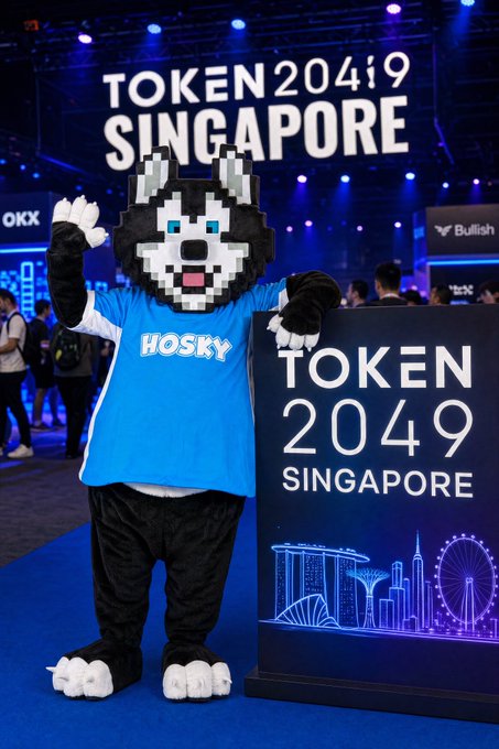 A person in a husky mascot costume wearing a blue 'HOSKY' t-shirt waves next to a 'TOKEN 2049 SINGAPORE' sign at a conference event.