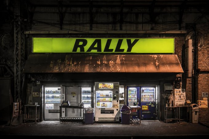 A nighttime view of a small shop with a bright yellow "RALLY" sign illuminates vending machines stocked with drinks, creating a striking urban scene. The image cleverly plays on the "Rally" name, evoking the idea of traders gathering at a late-night stop to fuel up—mirroring the post's tease of an upcoming announcement for the trading community.