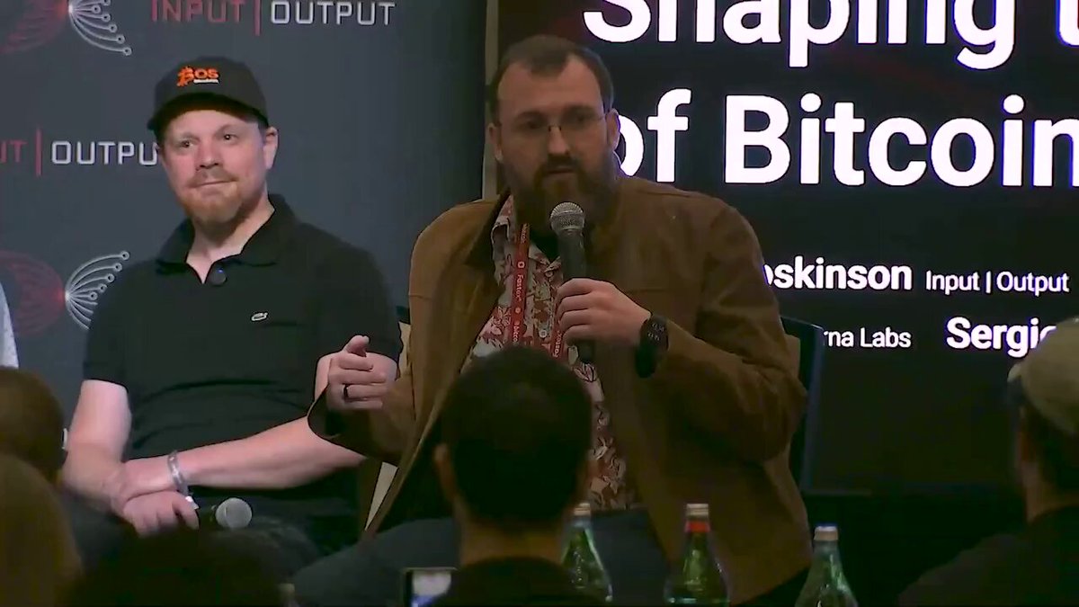 The image shows a man speaking into a microphone at an event with a large screen behind him displaying information about Bitcoin. The man appears to be discussing the shaping of Bitcoin, possibly as part of a panel discussion or presentation. In front of the speaker, there are two people sitting and listening attentively. On the right side of the image, there is a sign that says "The US government is going to hold  $ADA #Cardano". The background features a large screen displaying text related to Bitcoin.