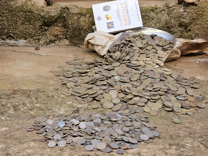 A metal bowl tipped over, spilling a large pile of old, tarnished coins onto a stone surface, accompanied by an information card for a 'SWANSEA CITY BITCOIN' token.