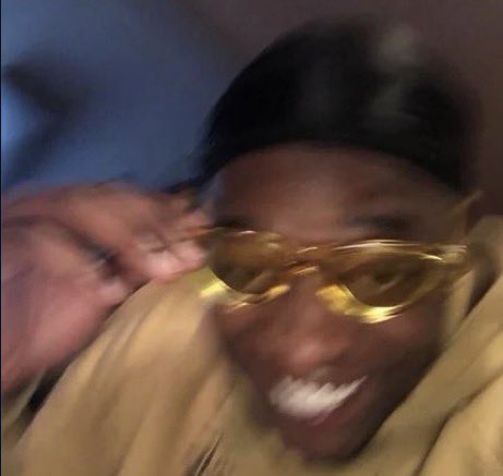 A blurry close-up photo shows a smiling person wearing yellow-tinted sunglasses and a beige hoodie, adjusting the frames with their hand.