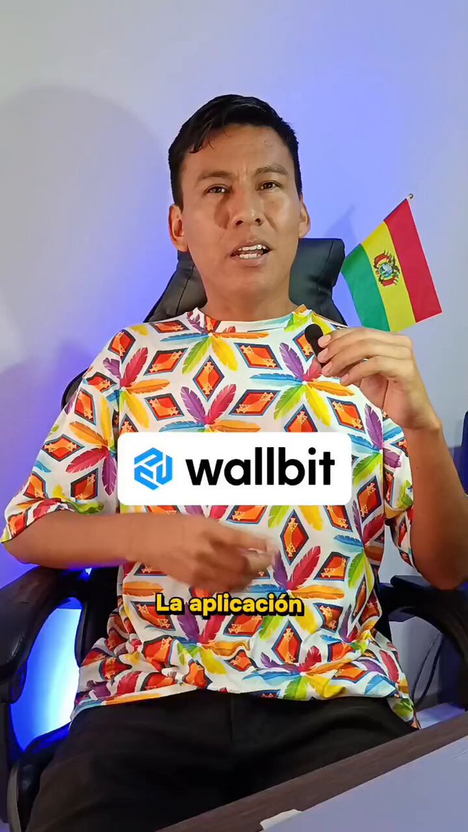 The image features a man sitting down, wearing a colorful shirt with a flag design on it. He is holding up a flag that has "Wallbit" written on it. The background of the image shows a computer screen displaying a message about an ETF called "Wallbit". This suggests that the man might be promoting or discussing Wallbit in some way.