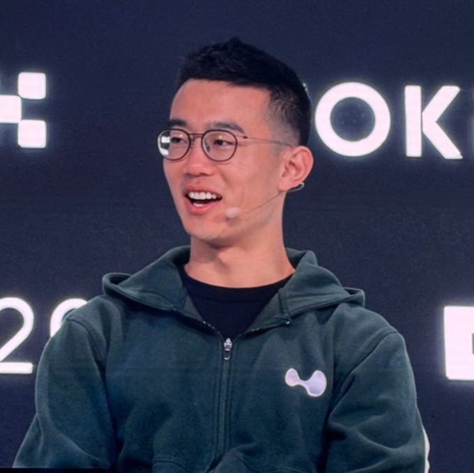 A man wearing glasses and a dark hoodie with a small logo speaks at an event, with 'OK' visible on the background screen.