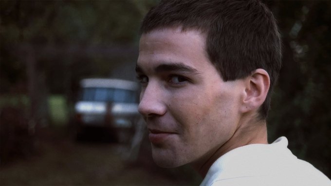 A young man with short dark hair glances over his shoulder with a knowing half-smile, an old vehicle blurred in the background. The image mirrors the post's message of skepticism, capturing the quiet amusement of someone who recognizes the disconnect between online hype and real results.
