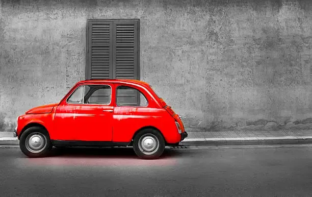 A vintage red Fiat 500 is parked on a quiet street in front of a weathered gray wall with a dark wooden shutter. The image humorously illustrates the "If fiat was a car:" caption by pairing the word "fiat" (a car brand) with its literal meaning—a decree or authorization—visually represented by a small, modest vehicle that could symbolize something officially sanctioned yet unimpressive in scale.