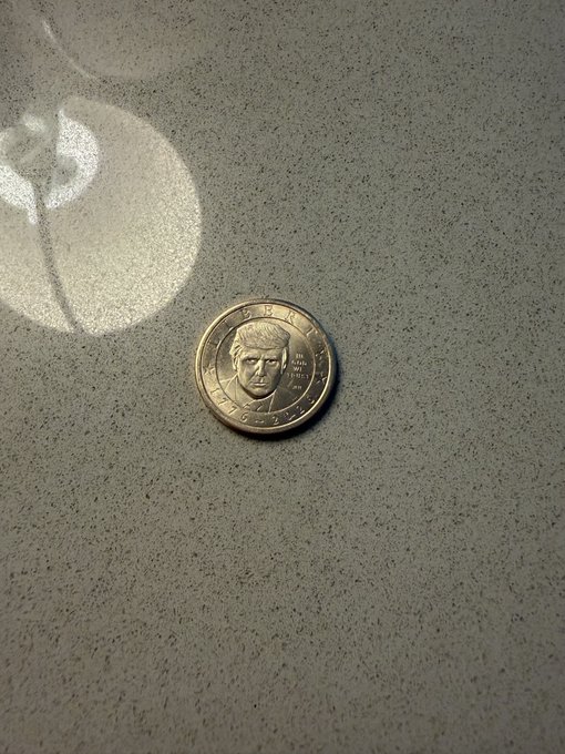 A gold-colored commemorative coin bearing Donald Trump's portrait and the inscription "TRUMP" rests on a speckled gray surface, matching the post's claim of a newly acquired Trump coin.