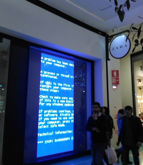 A digital billboard display inside a ZARA store, located near a "ZARA" storefront sign, shows a blue Windows Blue Screen of Death (BSOD) error message reading "A problem has been detected..." with technical information "*** STOP: 0x000000F4" while shoppers walk by carrying bags.