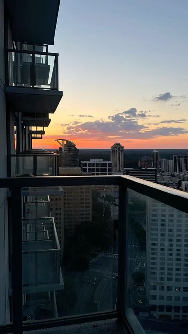 A view from a high-rise balcony overlooking a city skyline at sunset with orange and pink hues in the sky.