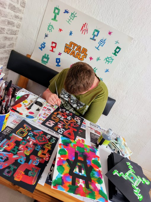 A person in a green shirt is seen from above working on colorful Star Wars-themed artwork at a table, with the official 'Star Wars' logo poster visible on the wall behind them and various vibrant paintings spread across the desk.